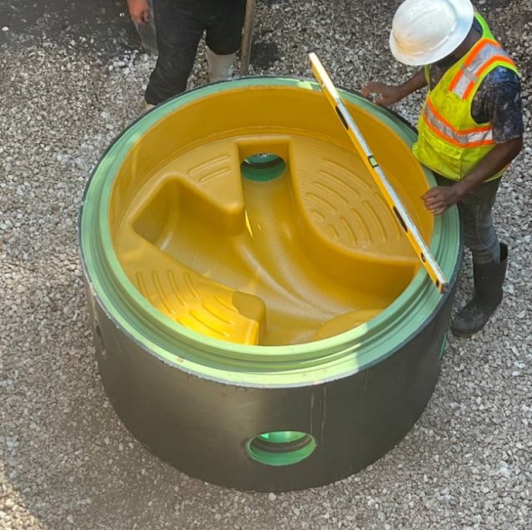 Sie sehen einen Schachtring mit Kunststoff-Schachtboden auf einer Asphaltboden. Daneben steht ein Abeiter und auf dem Schachtring liegt eine Wasserwage.